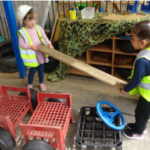 Two female pupils at Ormiston Herman Academy appear to be pretending to be builders, dressed up in high vis vests and construction helmets, are lifting a plank and surrounded by crates